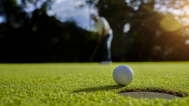 Golfçü topu yeşil golfün üstüne koyuyor, mercek akşam vakti parlıyor. Golfçü vuruşu uzun süre deliğe soktuktan sonra kazanır..                                