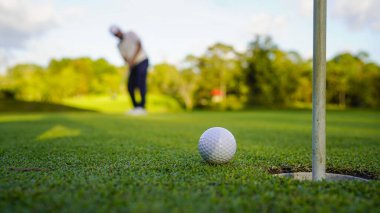 Golfçü topu yeşil golfün üstüne koyuyor, mercek akşam vakti parlıyor. Golfçü vuruşu uzun süre deliğe soktuktan sonra kazanır..                                