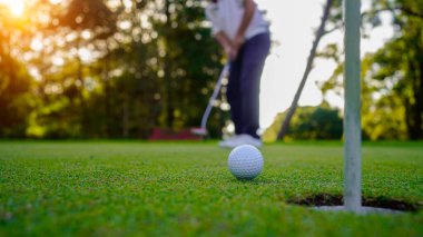 Golfçü topu yeşil golfün üstüne koyuyor, mercek akşam vakti parlıyor. Golfçü vuruşu uzun süre deliğe soktuktan sonra kazanır..                                 