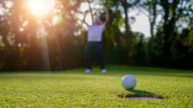 Golfçü topu yeşil golfün üstüne koyuyor, mercek akşam vakti parlıyor. Golfçü vuruşu uzun süre deliğe soktuktan sonra kazanır..                                 