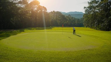 Golfçü topu yeşil golfün üstüne koyuyor, mercek akşam vakti parlıyor. Golfçü vuruşu uzun süre deliğe soktuktan sonra kazanır..                                