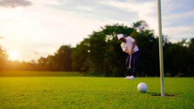 Golfçü topu yeşil golfün üstüne koyuyor, mercek akşam vakti parlıyor. Golfçü vuruşu uzun süre deliğe soktuktan sonra kazanır..                                 
