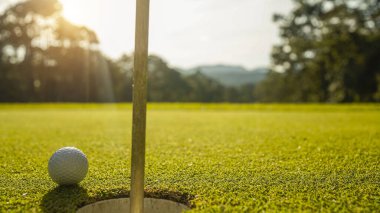 Golf topu yeşil çimlerin üzerinde, golf sahasında, arka planda güneş ışığı var. Yeşil çimlerin üzerindeki deliğin kenarındaki golf topu sıcak bir tonla ve günbatımıyla..                               