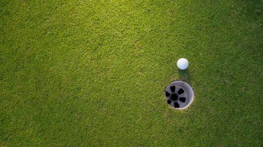 Golf topu yeşil çimlerin üzerinde, golf sahasında, arka planda güneş ışığı var. Yeşil çimlerin üzerindeki deliğin kenarındaki golf topu sıcak bir tonla ve günbatımıyla..                               