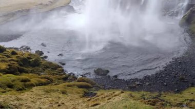 Yeşil yosun ve siyah çakıllarla çevrili bir şelale yere çarpar.