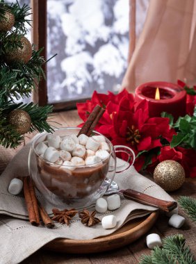 Bir fincan sıcak çikolata, marşmelovlu ve tarçınlı. Tatil süslemeleri ahşap bir masanın üzerinde. Kış arkaplanı olan bir pencerenin yanında.