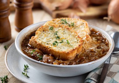 Fransız soğan çorbası, kızarmış peynirli baget ekmeği, kekik ile süslenmiş, ahşap bir masa arkasında.