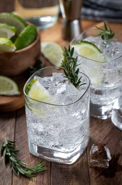 Gin and tonic cocktail with lime slices, rosemary and ice on a rustic wooden table