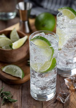 Gin and tonic cocktail with lime slices, and ice on a rustic wooden table