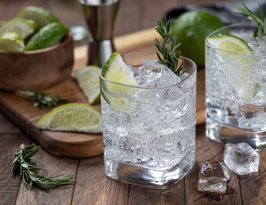 Gin and tonic cocktail with lime slices, rosemary and ice on a rustic wooden table
