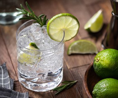 Gin and tonic cocktail with lime slices, rosemary and ice on a rustic wooden table