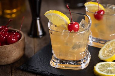 Whiskey sour cocktail with marachino cherry and lemon slice on black slate board