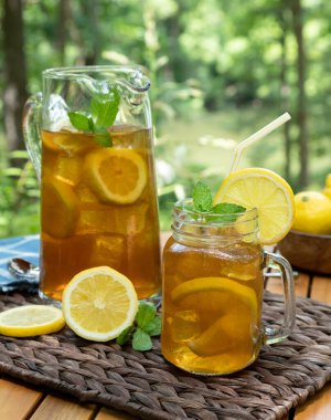 Cam kavanozda buzlu çay, limonlu sürahi ve açık havada nane, doğa arka planlı ahşap masada.