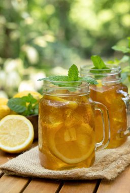 Limon dilimli cam kavanozlarda buzlu çay ve doğa arka planlı ahşap masada nane.