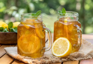 Limon dilimli cam kavanozlarda buzlu çay ve doğa arka planlı ahşap masada nane.