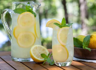 Limonata cam, sürahi ve naneli ve dilimlenmiş limonlar ahşap veranda masasında ve doğa arka planında.