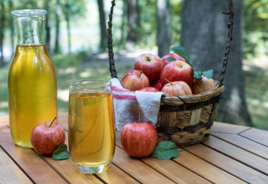 Elma suyu cam ve sürahide taze kırmızı elmalar ahşap teras masasında ve doğa arka planında.