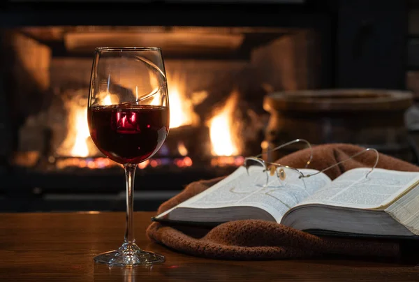 Glass of red wine with blanket and book on wooden table.  Burning fireplace in bqckground