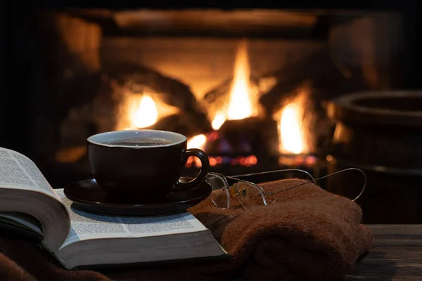 Cup of coffee with open book and blanket with a burning fireplace in background