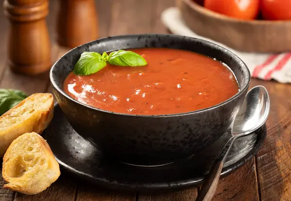 Fesleğen yapraklarıyla süslenmiş domates çorbası ve ahşap masada kızarmış baget dilimleri.