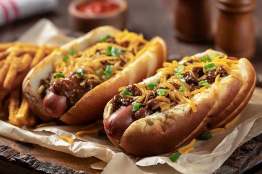 Two chili hot dogs with shredded cheddar cheese, chopped green onions and seasoned french fries on a wooden tray clipart
