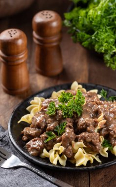 Siyah bir yemek tabağında maydanozla süslenmiş, yumurtalı erişteli biftek.