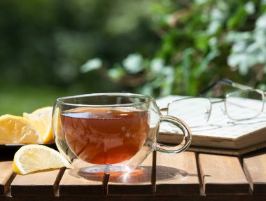 Bir fincan çay ve limon dilimleri ahşap masa üzerinde gözlüklü ve doğa arka planlı kitap.