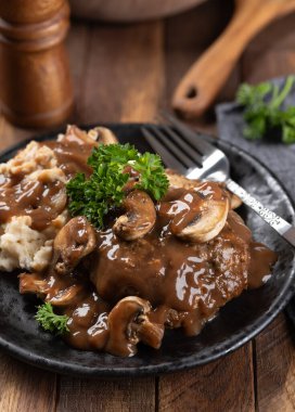 Maydanozla süslenmiş mantar soslu Salisbury bifteği ve siyah tabakta patates püresi.