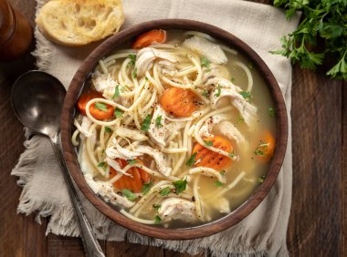 Bir kase tavuklu şehriye çorbası, peçetede havuç ve tahta masada baget dilimleri. Genel Görünüm