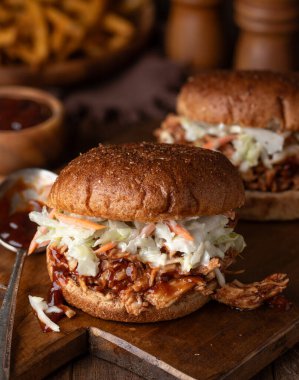 Barbekü tavuğu ve lahana salatalı sandviçi ahşap kesme tahtasının üzerinde, arka planda patates kızartması ile birlikte kepekli ekmeğin üzerine koydu.