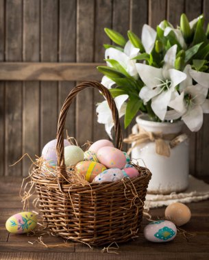 Sepette renkli Paskalya yumurtaları ve ahşap masa ve arka planda beyaz zambaklar.
