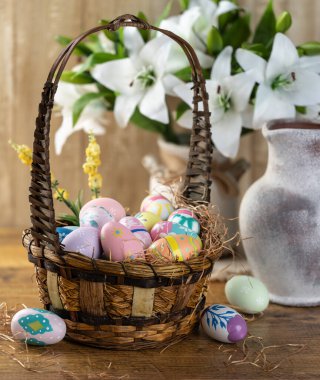 Paskalya yumurtaları beyaz zambaklarla süslenmiş bir sepette, ahşap arka planda.