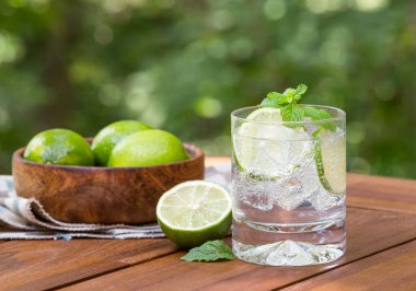 Bir bardak kireç dilimleri ve nane şekeri ile köpüklü su ve ahşap masa üzerinde doğa arka planlı yapraklar.
