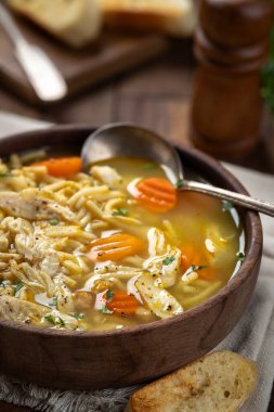 Bir kase tavuklu şehriye çorbası, peçetede havuç ve tahta masada baget dilimleri.