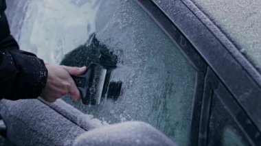 Kışlık ceketli bir adam, sıfırın altında bir kış sabahı, pencerelerindeki ağır don ve buzları ve donmuş bir arabanın ön camını sökmek için bir buz kazıyıcı kullanır..