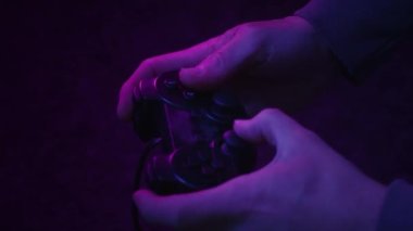 Bir bilgisayar oyuncusu, sadece pembe ve mavi titreşimli bir TV ekranı tarafından aydınlatılan markasız bir oyun denetleyicisinin düğmelerindeki parmak çubuklarını ve çekiçleri hızla kaydırır..