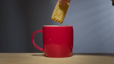 Bir adamın eli bisküviyi sıcak bir içeceğe bandırır parlak kırmızı bir kahve bardağında..