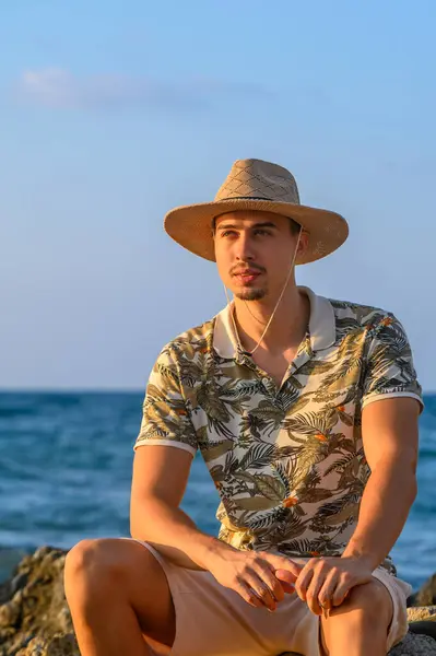 a young guy with a mustache and a hat sits on a stone on the shore of the Mediterranean Sea 11