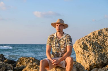 Akdeniz kıyısında bir taşın üzerinde bıyıklı ve şapkalı genç bir adam oturuyor.