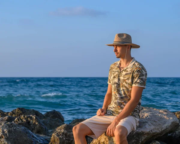 a young guy with a mustache and a hat sits on a stone on the shore of the Mediterranean Sea 6