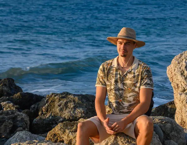 a young guy with a mustache and a hat sits on a stone on the shore of the Mediterranean Sea 1