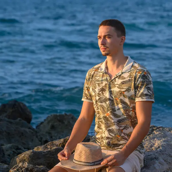 a young guy with a mustache sits on a stone on the shore of the Mediterranean Sea