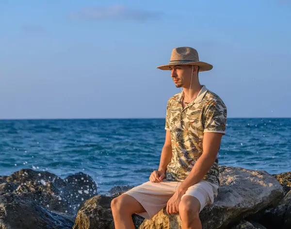 a young guy with a mustache and a hat sits on a stone on the shore of the Mediterranean Sea 9