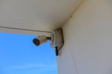 A surveillance camera discreetly installed at a corner captures the endless blue sky, quietly observing the surroundings while blending into the architecture. clipart