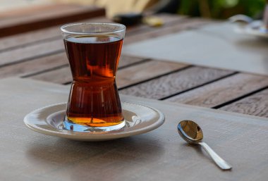 Altın kahverengimsi çay eşsiz şekilli bir bardağı doldurur, gümüş kaşığın yanındaki bir çay tabağının üzerinde dinlenir, hepsi de rustik ahşap masalar ve yemyeşil yemyeşil bir zemin üzerine kuruludur..