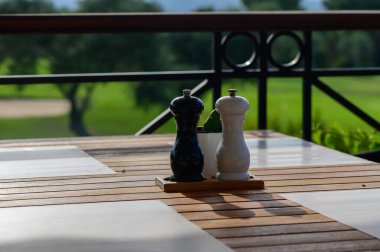 A serene outdoor dining area showcases elegant black and white pepper shakers resting on a polished wooden table, blending with vibrant greenery in the background. clipart