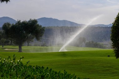 Güneş ufuktan yükselirken fıskiyeler canlı yeşil golf sahasını hafifçe buğulandırarak sakin bir atmosfer yaratıyor. Uzak tepeler heybetli duruyor, dingin güzelliği arttırıyor..