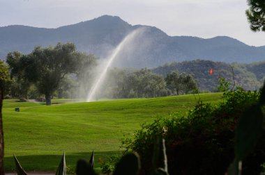 Sabah ışığında sakin bir golf sahası canlanır, yemyeşil çimenler ve sulama sisteminden fışkıran köpüklü su, görkemli dağlara çarpar..