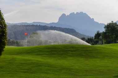 Serinletici su, uzak dağların ihtişamlı bir şekilde arka planda durduğu canlı golf sahası boyunca hafifçe püskürtür, sakin bir sabah titreşimini yakalar..