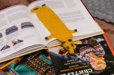 A handmade yellow knitted bookmark in the shape of an animal lies over two open books filled with colorful textile patterns and knitting techniques, inviting creativity. clipart